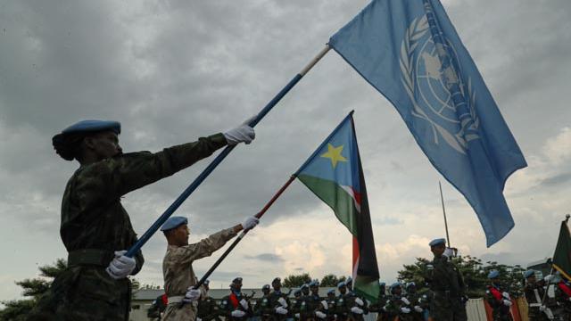 Two peacekeepers each holding a flag. One is holding the UN flag and the other is holding the South Sudanese flag.